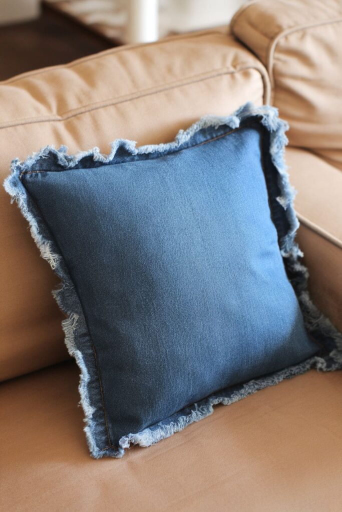 Casual, frayed-edge denim pillow with two denim squares glued together, leaving one side open for stuffing, giving it a relaxed, lived-in look