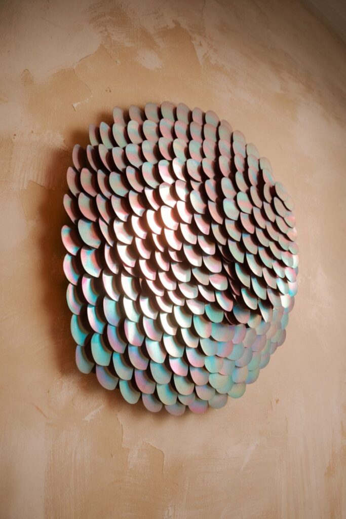 Wall art with overlapping crescent-shaped cardboard pieces resembling fish scales, painted in shimmering ombre colors