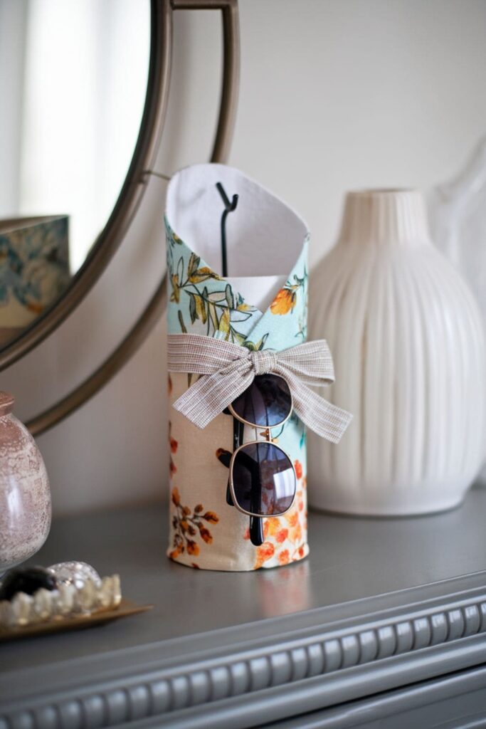 A DIY sunglasses holder made from a decorated paper towel roll, featuring a small hook inside to hang sunglasses on a chic dresser