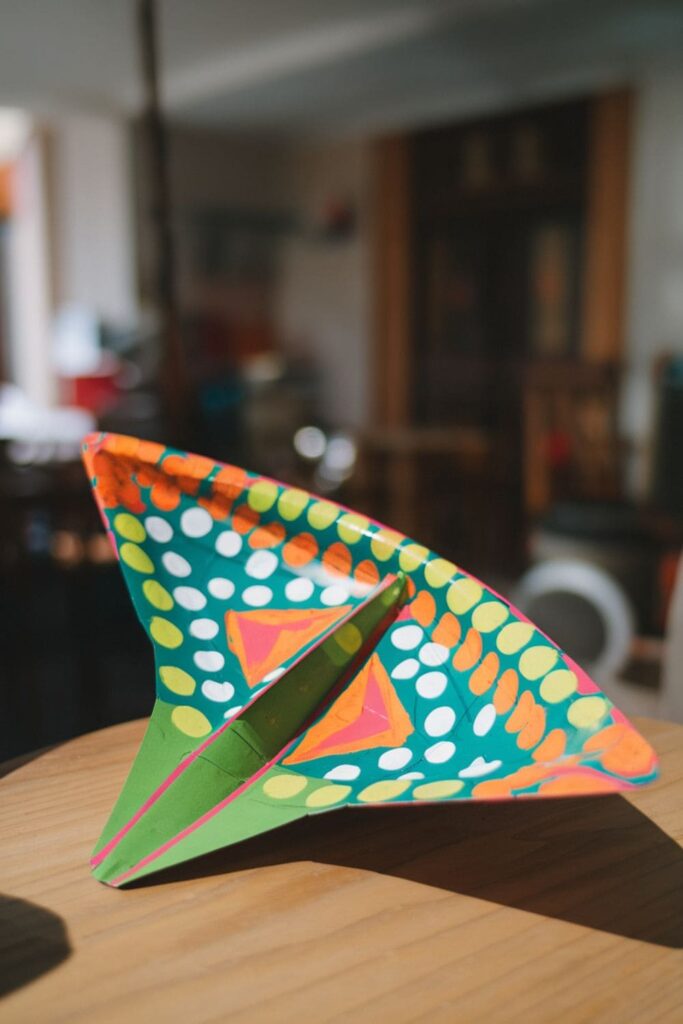 A brightly painted paper plate designed as a flying disc with vibrant patterns, shown outdoors mid-flight