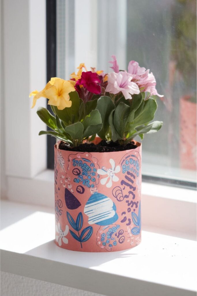 A flower pot created from a decorated paper towel roll, filled with soil and blooming flowers, placed on a sunny windowsill