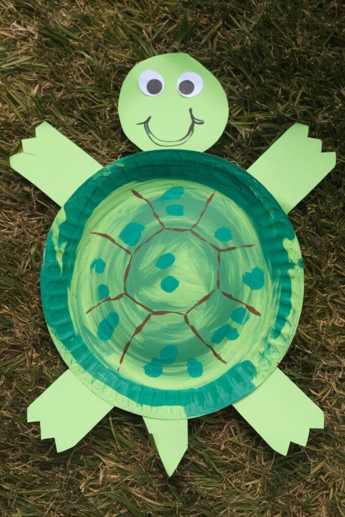 A green-painted paper plate turned into a turtle, with added construction paper legs, head, and tail, placed on a grassy surface