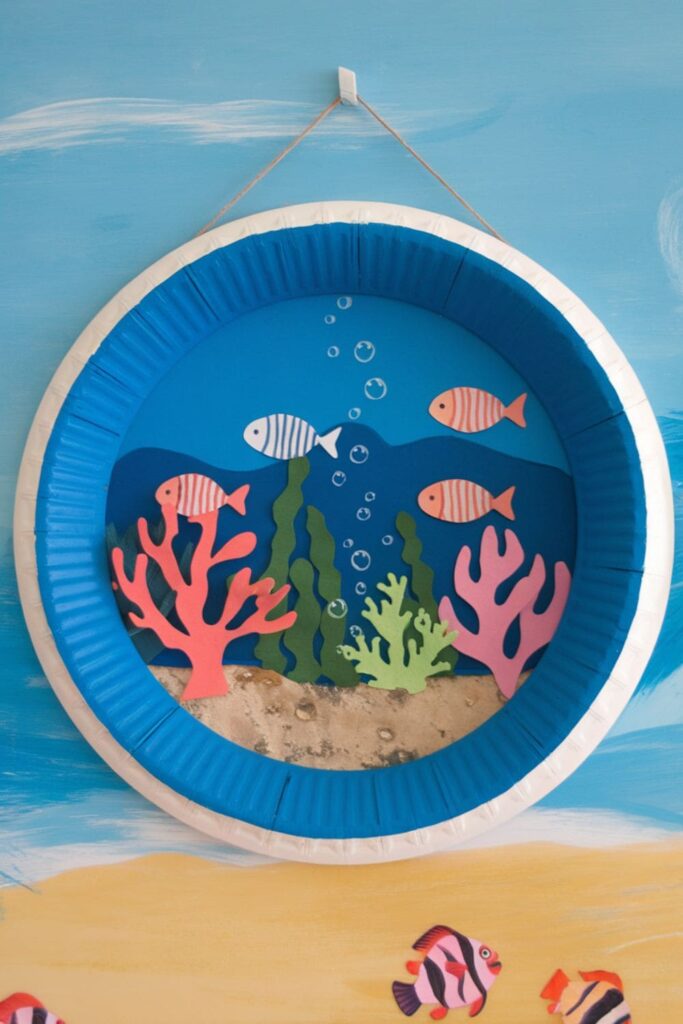 A paper plate painted blue with cutouts of fish, coral, and seaweed, resembling an underwater porthole