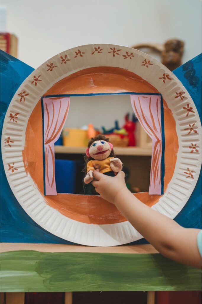 A small puppet theater made from a paper plate with a cut-out window, painted and decorated with stars and curtains