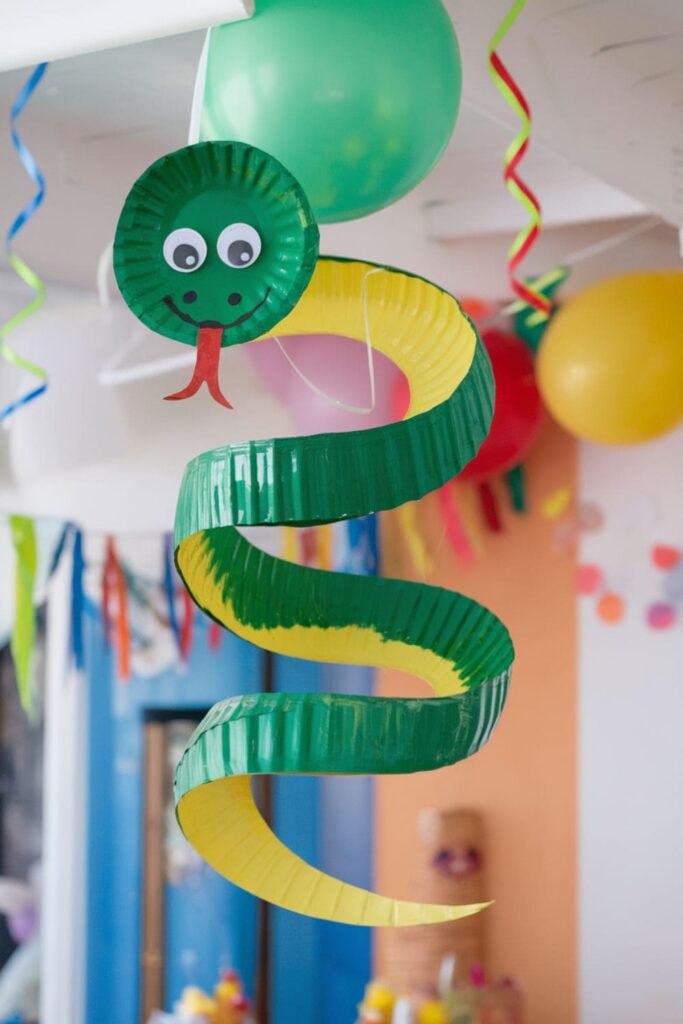 A spiral-cut paper plate painted with green scales, featuring googly eyes and a red forked tongue, hanging from the ceiling