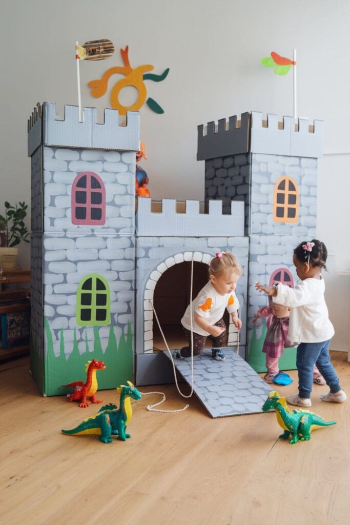 Cardboard castle with crenellated walls, a functional drawbridge, gray stone-painted texture, and colorful cutout windows, surrounded by kids playing