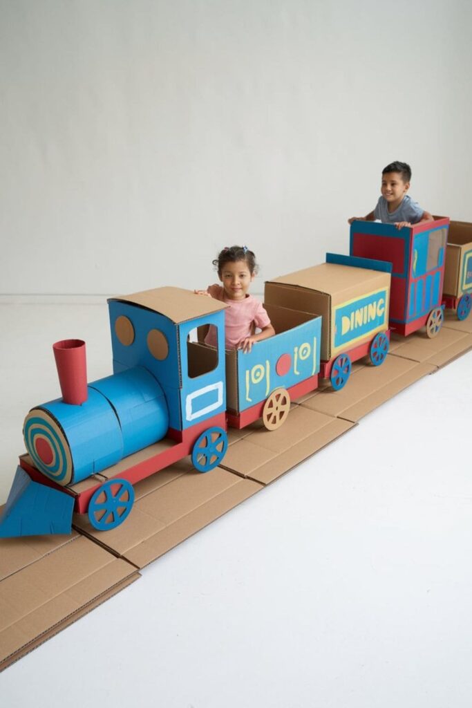 Cardboard train set with multiple connected boxes painted in different colors, a cardboard track beneath, and children pretending to ride the train