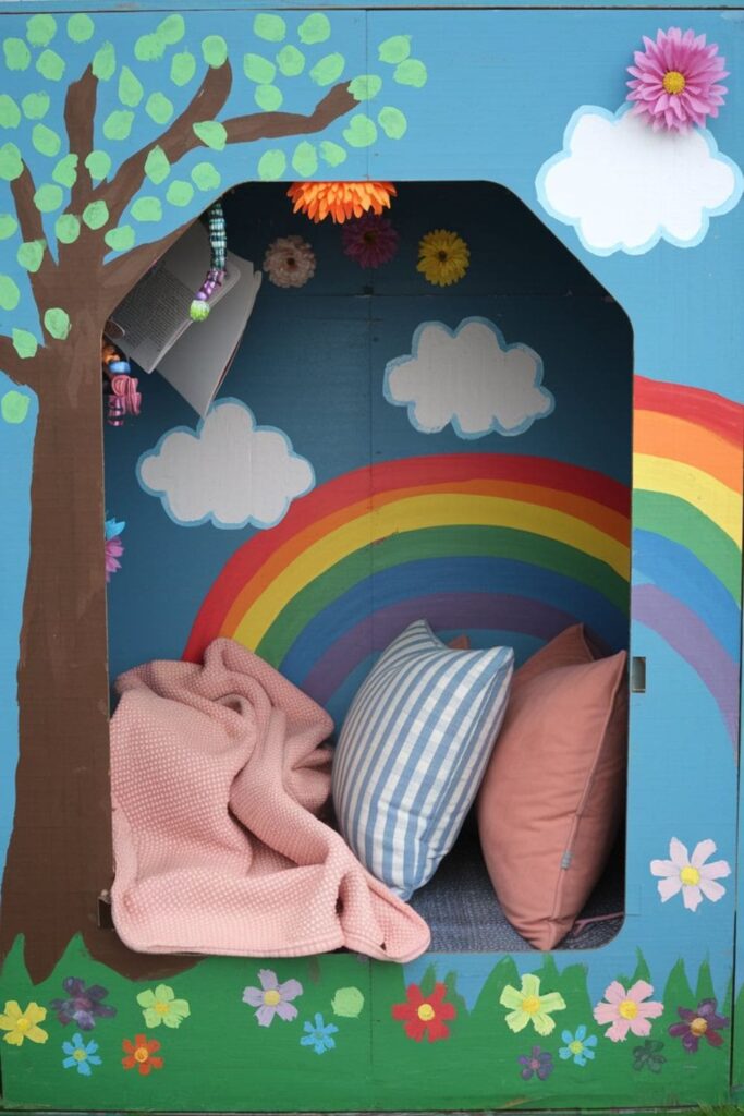 Cozy cardboard reading nook decorated with stickers, painted designs, artificial flowers, and a soft interior with pillows and a blanket