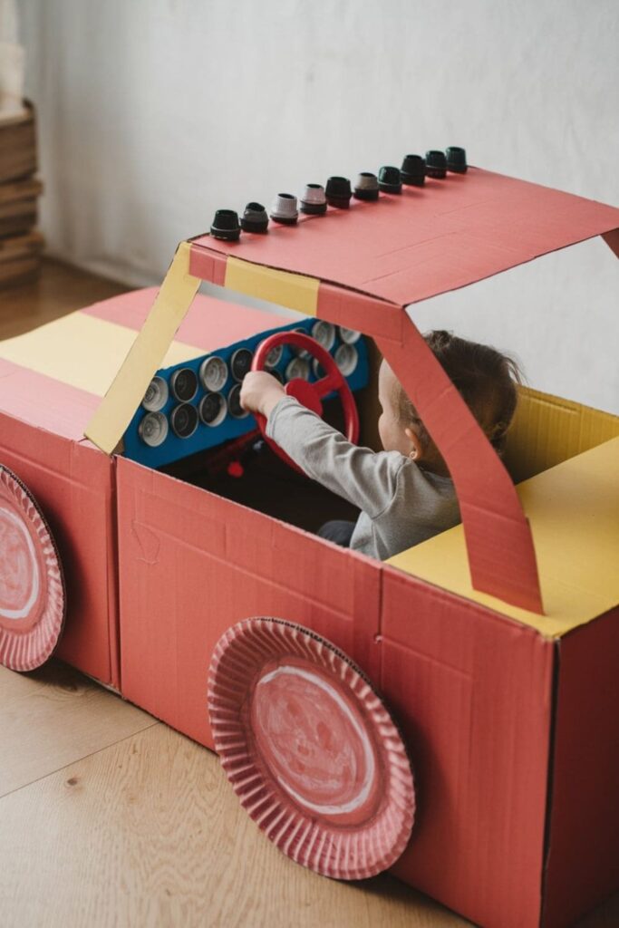 DIY cardboard race car painted red and yellow, with paper plate wheels, a bottle cap dashboard, and a child sitting inside pretending to drive