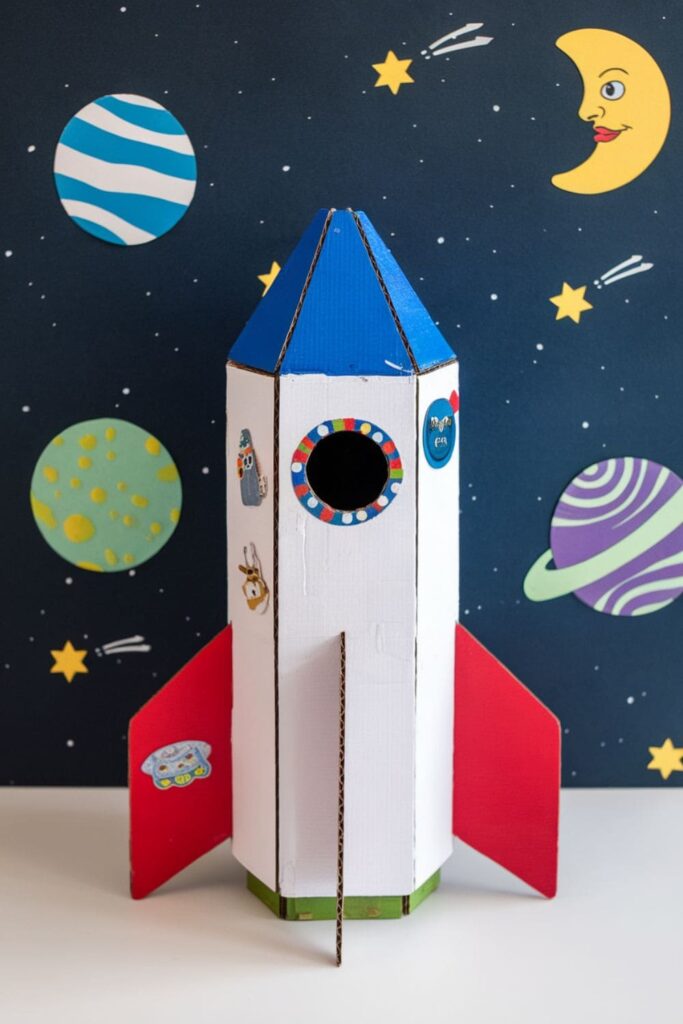 Handmade rocket ship crafted from a cardboard roll, painted white with red fins and a cone-shaped top, set against a starry background