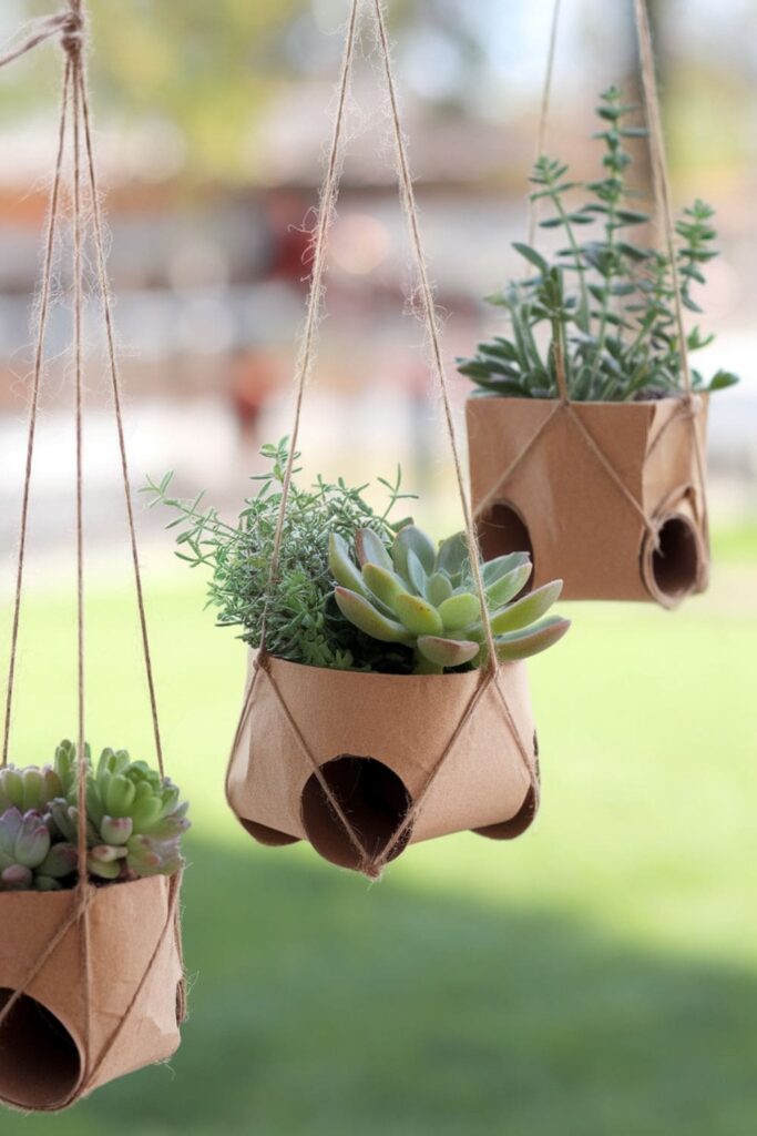 Hanging planters made from cut paper towel rolls, filled with small succulents and herbs, suspended by twine against a bright outdoor background