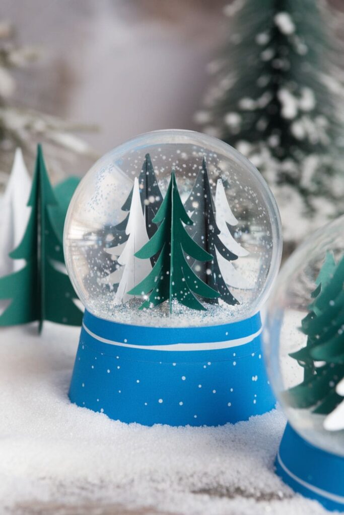 Whimsical paper snow globe created from a clear plastic cup, filled with cut-out paper trees and glitter, set against a snowy background